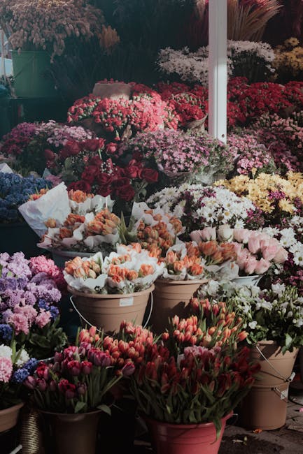 A vibrant display of various fresh flowers, including pink, red, peach, purple, and white blooms, arranged in brown, beige, and pink pots and buckets at a flower stall. The arrangement features tulips
