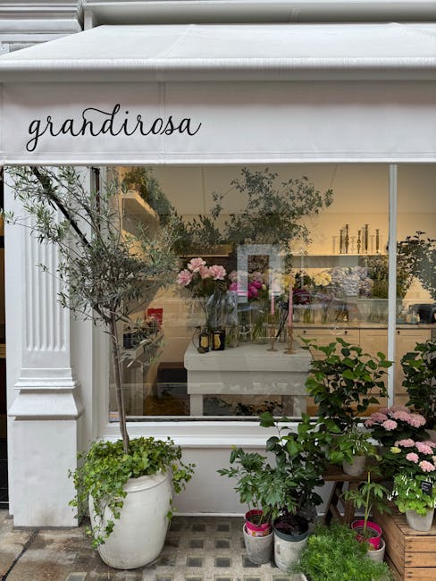 The image shows the exterior of Grandiroza, a florist shop with a white storefront and large glass window displaying a variety of fresh floral arrangements inside. Outside the shop, there are potted p