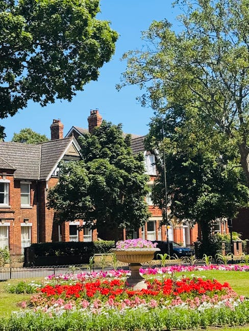 The image depicts a vibrant, well-maintained flowerbed in a public park, featuring a colorful arrangement of red, pink, and white flowers in full bloom, set against a backdrop of lush green trees and