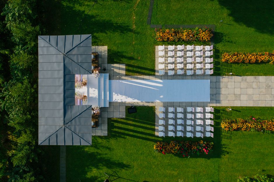 An aerial view of an outdoor wedding setup featuring a rectangular aisle covered with a white runner, leading to a floral arch decorated with soft pink and white roses, peonies, and greenery. Flanking