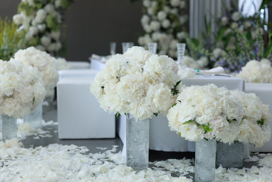 A floral arrangement featuring large, rounded bouquets of white hydrangeas and peonies placed in tall, rectangular concrete vases. The flowers are fresh, with soft, layered petals displaying a delicat